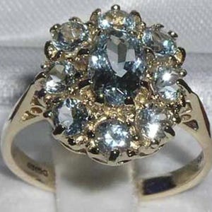 May include: A gold ring featuring an oval, light blue gemstone surrounded by smaller, round, light blue gemstones. The ring's band is gold with intricate detailing. The gemstones are set in a cluster design.