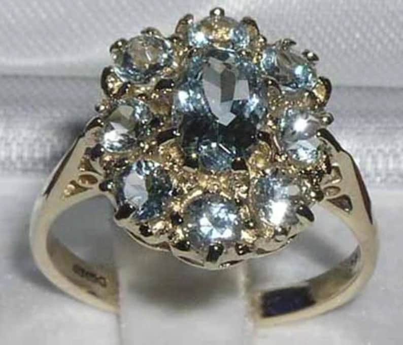 May include: A gold ring featuring an oval, light blue gemstone surrounded by smaller, round, light blue gemstones. The ring's band is gold with intricate detailing. The gemstones are set in a cluster design.