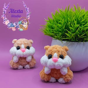 May include: Two handmade crochet hamster plushies. The plushies are light brown with white and pink accents. The hamsters have black eyes and are sitting on a purple surface. A green plant in a white pot is in the background.