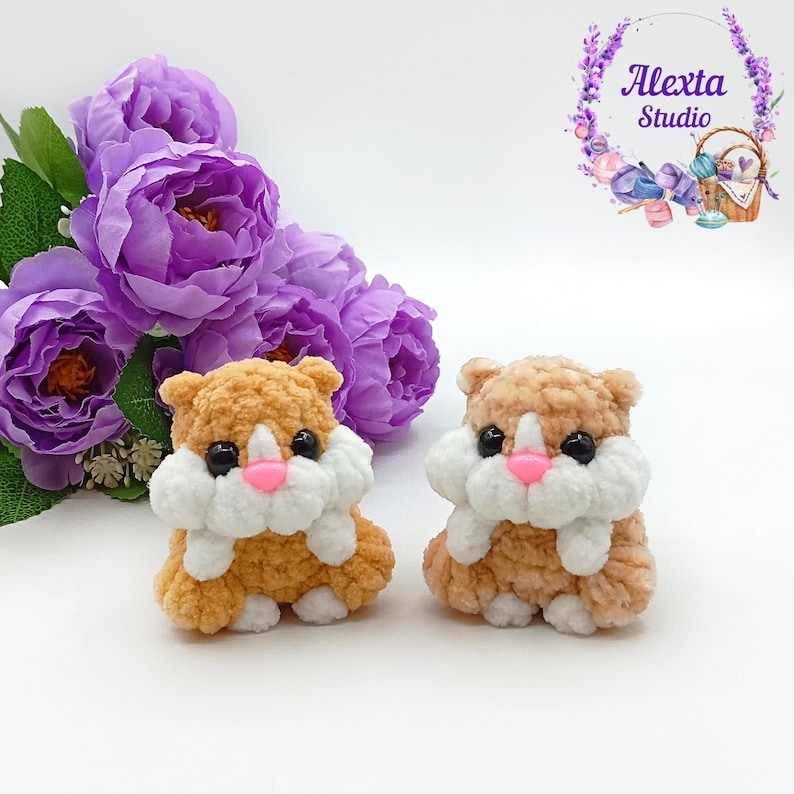 May include: Two handmade crochet hamster toys in shades of brown and white, with black eyes and pink noses. The toys are sitting on a white surface, with purple flowers in the background. The toys are from Alexta Studio.