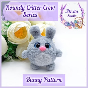 Może przedstawiać: Pluszowy szary królik z dużymi niebieskimi oczami i różowym nosem. Królik jest częścią "Roundy Critter Crew Series" i jest oznaczony jako "Bunny Pattern". Tło zawiera białe kwiaty i logo "Alexta Studio".