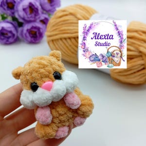 May include: A handmade, plush hamster toy in shades of brown, white, and pink. The hamster has black eyes and a pink nose and paws. The image also shows a ball of yarn and a logo that says "Alexta Studio".