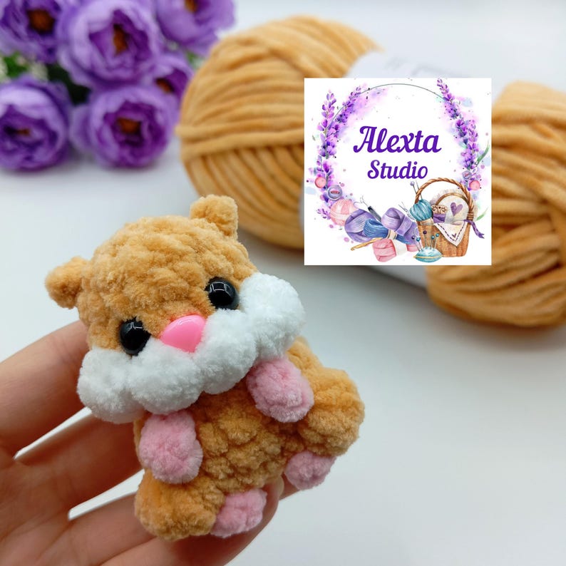 May include: A handmade, plush hamster toy in shades of brown, white, and pink. The hamster has black eyes and a pink nose and paws. The image also shows a ball of yarn and a logo that says "Alexta Studio".