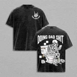 May include: A black acid-wash t-shirt featuring a white skeleton hand graphic on the front with the text COOL DADS CLUB. The back displays a skeleton sitting on a toilet looking at a phone with the text DOING DAD SHIT and a roll of DAD TISSUES.