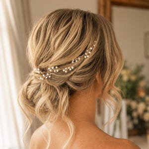 May include: A woman's blonde hair styled in an updo, decorated with a delicate gold hair vine with small, white pearls. The hair accessory adds elegance to the hairstyle, suitable for weddings or special occasions.