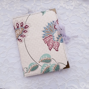 Op de afbeelding: Een witte stoffen journalhoes met een bloemenpatroon in tinten roze, blauw en bruin. De hoes heeft een decoratieve kanten rand en een gouden metalen hoek.