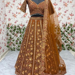 May include: A brown, intricately embroidered lehenga choli set. The set includes a short-sleeved top and a full skirt, both adorned with gold floral patterns and embellishments. A matching sheer dupatta with detailed borders completes the ensemble. The text "Premuati Ethnic" is visible.