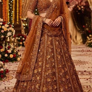 May include: A brown, embellished lehenga choli set. The skirt and short-sleeved top are covered in gold floral embroidery and sequins. A matching sheer dupatta is draped over the shoulders. The outfit is worn with a matching necklace and bracelets.