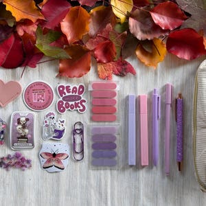 May include: An assortment of stationery items in shades of pink and purple, including pens, highlighters, and stickers, alongside a pencil case. The items are arranged on a light surface, with a backdrop of autumn leaves. The stickers have text that reads "Read More Books".