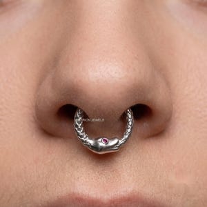 May include: A silver septum ring shaped like a snake biting its own tail, featuring a textured scale pattern along the body and a small, round, pink gemstone set into the head. The text NEUTRION JEWELS is visible on the metal surface.