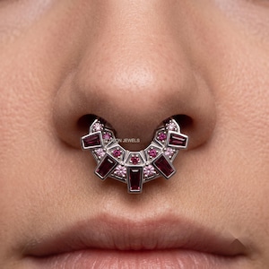 Könnte beinhalten: Ein silberner Septum-Ring mit halbkreisförmigem Design, verziert mit mehreren tiefroten Baguette-Schliff-Steinen und kleineren runden rosa Akzentsteinen. Die Metallfassung ist poliert und trägt mittig den Text VELTRION JEWELS.