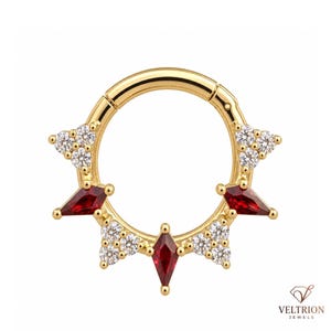 May include: A gold hinged hoop ring featuring a decorative sunburst design. The piece is adorned with three kite-shaped red gemstones and four clusters of three small clear round stones. The text VELTRION JEWELS is visible in the bottom right corner.