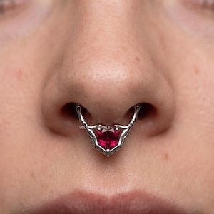 May include: A silver-toned septum ring featuring two skeletal hands meeting at the centre to hold a vibrant, heart-shaped red gemstone. The jewellery is positioned in the nasal septum, with the text VELVETUN JEWELS visible on the metal surface.