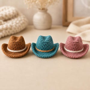 以下が含まれることがあります： 茶色、ティール、ダスティピンクの糸で編まれた3つのミニチュアのカウボーイハット。各ハットは、つまんだクラウン、カーブしたつば、クラウンのベースを囲む細い編み込みバンドが特徴です。茶色とピンクのハットには白いバンドが付いています。