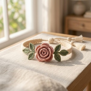 Puede incluir: Una diadema de ganchillo color crema con una rosa rosa y hojas verdes. La diadema tiene un diseño trenzado y borlas. El diseño floral es el punto focal, probablemente para el cabello.