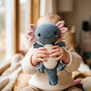 Peut inclure: Une peluche axolotl au crochet faite à la main, de couleurs bleu, rose et crème. L'axolotl a un visage souriant, des branchies frangées et de petits membres. La peluche est tenue par une personne portant un pull blanc.