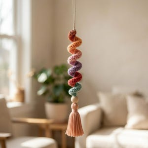 Pode incluir: Decoração suspensa em crochê em forma de espiral em cores do arco-íris, incluindo laranja, roxo, vermelho e verde. Uma conta de madeira e uma borla cor de pêssego são presas na parte inferior. A decoração é suspensa por um fio fino de cor natural.