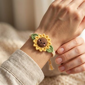 May include: A handmade crochet bracelet featuring a smiling sunflower design. The sunflower has yellow petals, a brown centre, and green leaves. The bracelet is cream-coloured with a yellow tassel.