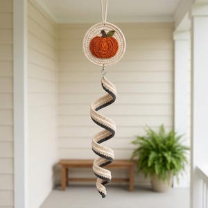 Puede incluir: Un adorno colgante de ganchillo con un disco circular superior que presenta una aplicación de calabaza naranja con una hoja verde. Debajo del disco, cuelga una larga cola en espiral hecha de rayas alternas de color crema y gris oscuro.