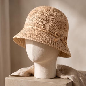 May include: A light tan, woven bucket hat with a rounded crown and a downward-sloping brim. The hat features a braided band encircling the base of the crown, finished with a small, tied bow detail on the right side. The texture is a tight, uniform knit.
