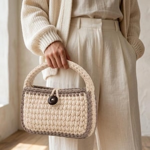 May include: A rectangular crochet handbag featuring a cream-colored body with a thick taupe border and a matching rounded handle. The bag is secured with a single dark brown circular button and a loop closure, showcasing a textured yarn construction.