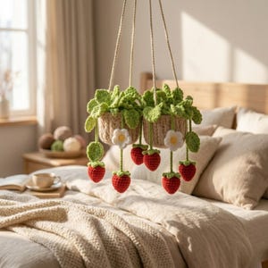 Può includere: Due fioriere sospese all'uncinetto con foglie verdi, fiori bianchi e fragole rosse. Le fioriere sono sospese da cordoncini beige. La scena è ambientata in una camera da letto con un letto e una finestra sullo sfondo.