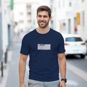 Op de afbeelding: Marineblauw T-shirt met een nep Windows-foutmelding met de tekst "Your code works on the first try." Het T-shirt heeft een klassieke ronde hals en wordt door een persoon gedragen.
