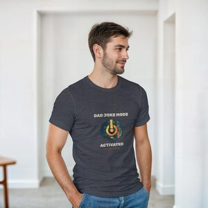 May include: Dark gray t-shirt with the text "DAD JOKE MODE ACTIVATED" and a power button graphic. The graphic is a circle with blue, yellow, and orange colors. The shirt is short-sleeved and made of a soft material.