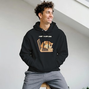 Puede incluir: Sudadera con capucha negra con el texto "I WORK IN COMPUTERS" sobre un gráfico de un gato atigrado naranja durmiendo con auriculares en una computadora portátil. Una taza de café está al lado de la computadora portátil.
