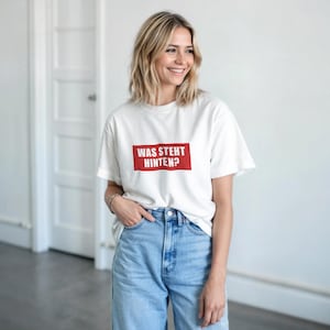Puede incluir: Camiseta blanca con el texto alemán "WAS STEHT HINTEN?" en un rectángulo rojo. La camiseta se combina con vaqueros azul claro. La modelo sonríe.