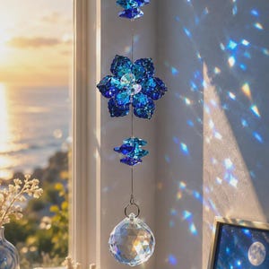 May include: A hanging suncatcher featuring a faceted clear crystal sphere at the bottom, connected by a thin wire to a deep blue crystal flower and two smaller blue crystal accents. The crystals cast light reflections on the wall.