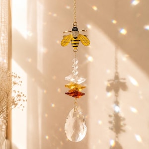 May include: A hanging suncatcher ornament featuring a gold-toned metal bee with yellow and black enamel wings and a crystal-encrusted head. Below the bee are three stacked geometric crystals in clear, amber, and brown, ending in a clear teardrop prism.