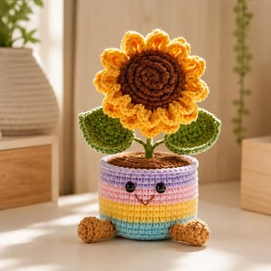 May include: A crocheted sunflower with a dark brown center and bright yellow petals, featuring two green leaves on a stem. The flower is planted in a pastel-striped pot with a smiling face, black eyes, and two small brown feet at the base.