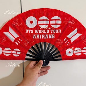 Pode incluir: Um leque dobrável vermelho com varetas pretas apresentando o texto BTS WORLD TOUR ARIRANG em branco. O leque exibe logotipos BTS brancos, padrões gráficos circulares e várias assinaturas impressas por toda a superfície.