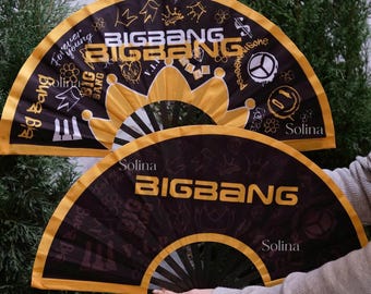 Op de afbeelding: Twee zwarte en gele opvouwbare handwaaiers met de tekst BIGBANG in vetgedrukte letters. De waaiers zijn versierd met diverse witte en gouden grafische iconen, waaronder kronen, vredestekens en muzieknoten, samen met de merknaam Solina.