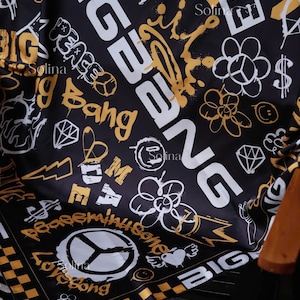 Könnte beinhalten: Ein schwarzes quadratisches Tuch mit einem gold-weißen Graffiti-Druck. Das Design enthält den Text BIGBANG, PEACEMINUSONE und LOVESONG sowie Symbole wie Friedenszeichen, Smileys, Diamanten, Blumen und einen karierten Rand.