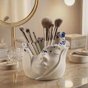 Puede incluir: Un porta-brochas de maquillaje de cerámica blanca con forma de rostro, con detalles de salpicaduras azules. El soporte contiene varios pinceles de maquillaje. Se encuentra sobre una encimera de mármol, con un espejo y otros accesorios de belleza en el fondo.