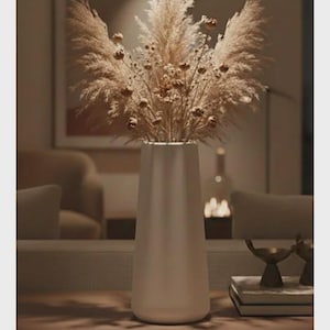 May include: A white vase, holding dried pampas grass and small dried flowers, stands on a wooden surface. The background is blurred, showing a living room. The colour scheme is neutral and warm, creating a calming atmosphere.