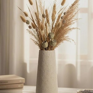 May include: A beige textured vase filled with dried wheat, pampas grass, and seed pods. The vase has a conical shape and sits on a light-colored wooden surface. The arrangement is a neutral-toned home decor accent.