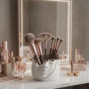 May include: A white ceramic face-shaped makeup brush holder filled with various makeup brushes. The brushes have brown and rose gold handles. The holder sits on a marble vanity with a lighted mirror in the background. Other makeup items are also visible.