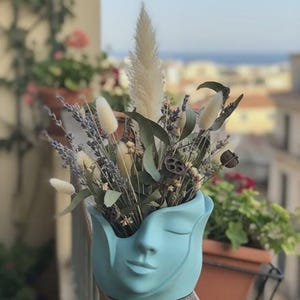 May include: A pale blue ceramic vase in the shape of a face, holding dried flowers and foliage. The arrangement features lavender, pampas grass, and other natural elements, creating a tranquil and artistic display. The vase is placed on a wooden railing.