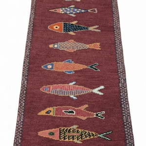 May include: A long, rectangular runner rug in a deep maroon color featuring a vertical row of eleven stylized fish motifs. Each fish is rendered in a unique combination of colors including orange, blue, yellow, and green, with a patterned border.