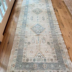 May include: A long, rectangular runner rug featuring a cream-colored base with a wide, muted blue border. The central field displays a series of repeating diamond-shaped medallions in soft blue and tan tones, accented by small, intricate geometric motifs.
