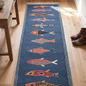 May include: A long, rectangular blue runner rug featuring a repeating pattern of stylized fish in various colors including orange, pink, and yellow. The rug has a decorative geometric border and white fringe at both ends, laid out on a wooden floor.