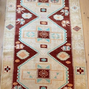 May include: A rectangular area rug featuring a central geometric pattern with large diamonds and cross motifs. The design incorporates deep red, cream, light blue, and soft orange colors, with repeating floral-like shapes along the borders.