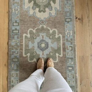 May include: A rectangular runner rug featuring a muted brown base with two large, symmetrical floral-inspired medallions in shades of sage green, light blue, and cream. The rug is framed by a decorative border with geometric patterns.