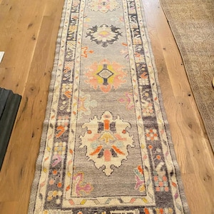 May include: A long, narrow runner rug featuring a gray base with a repeating series of intricate, symmetrical floral medallions in shades of coral, yellow, light blue, and dark charcoal. The border displays a geometric pattern in matching colors.