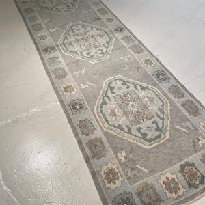 May include: A long, rectangular runner rug featuring a light gray base with three prominent central geometric medallions in shades of sage green, soft blue, and beige. The border is decorated with repeating square floral motifs in muted tones.