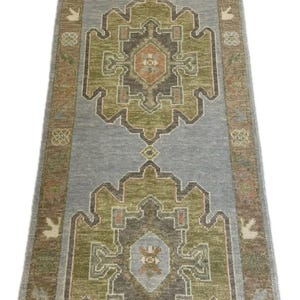 May include: A long runner rug featuring a light blue background with two large, repeating geometric medallions in olive green, charcoal gray, and muted orange. The border displays a repeating pattern of small floral motifs in brown and tan.