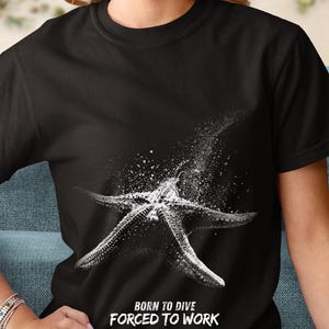 Può includere: Una t-shirt nera a maniche corte con una grafica bianca di una stella marina con spruzzi di sabbia. Sotto la stella marina, il testo BORN TO DIVE FORCED TO WORK è stampato in un carattere bianco dall'aspetto usurato, centrato sul petto.